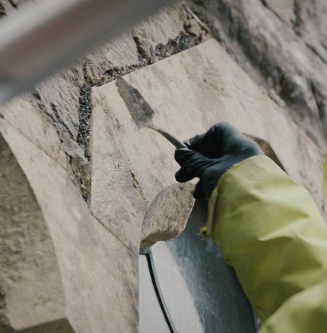 Skilled mason using a trowel to carefully repair and regrout the historic stone wall of a building.